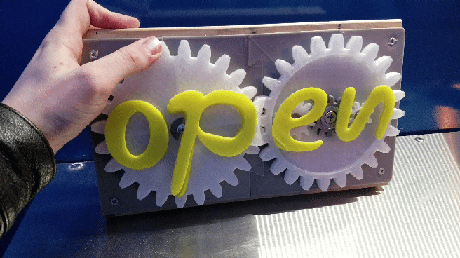 a 3d printed sign that consists of two gears with the letters "clo" on one gear and "sed" on the other. Animation starts with the gears showing the word "closed." A hand turns the gears, and the letters turn and overlap in such a way that the sign now reads "open"
